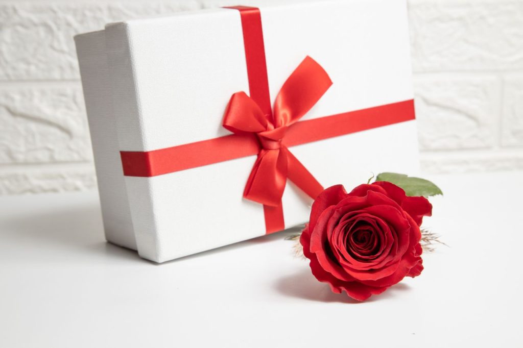 A white gift box with a red ribbon and a vibrant red rose, ideal for Valentines Day or special romantic occasions.